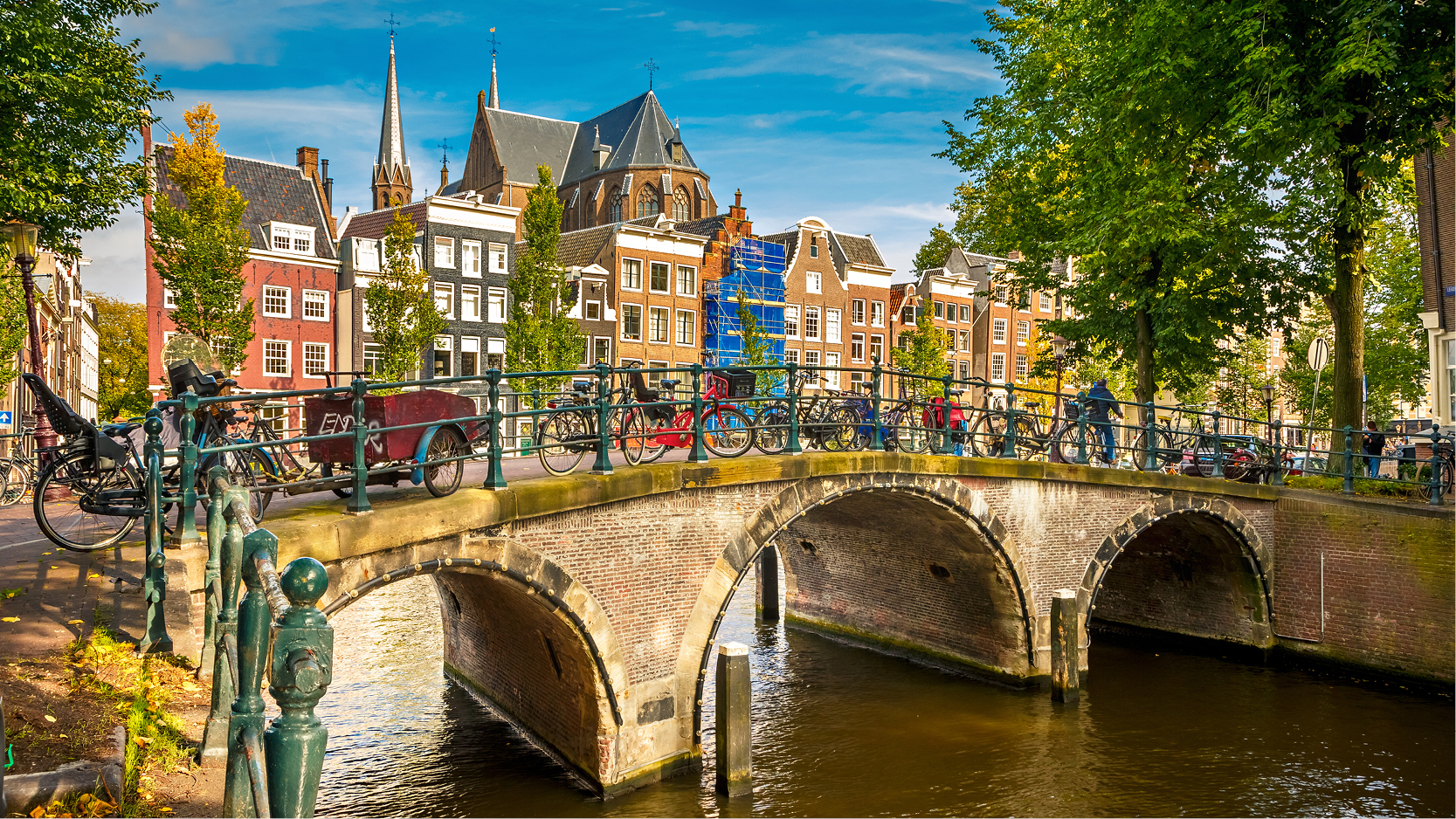 Kullerstensbro med cyklar ver kanalen i Amsterdam. hus och grna trd i bakgrunden.
